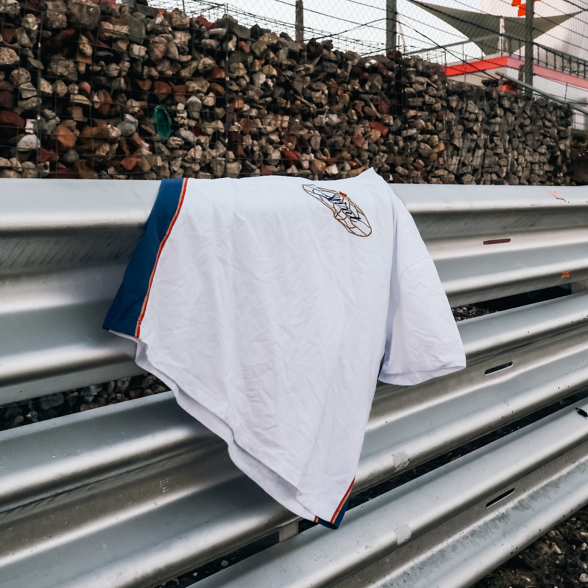962 Tee by STINT GEAR. Blue, gold and red 956 and 962 livery on white, draped over a crash barrier at the circuit. Motorsport livery apparel.