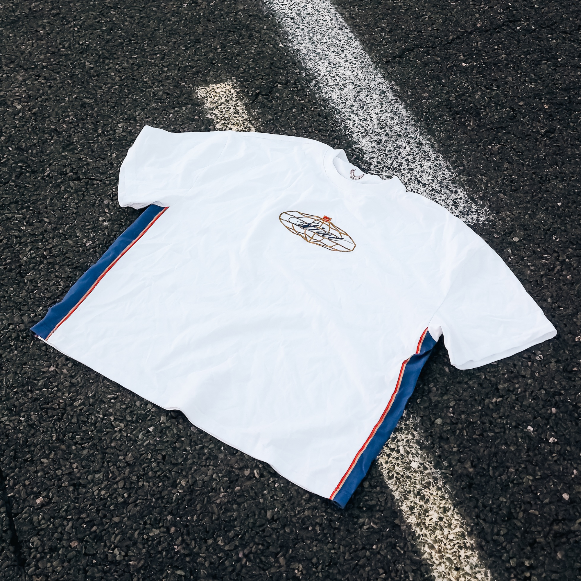 962 Tee by STINT GEAR. Blue, gold and red 956 and 962 livery on white, laid on painted track asphalt. Motorsport livery apparel.