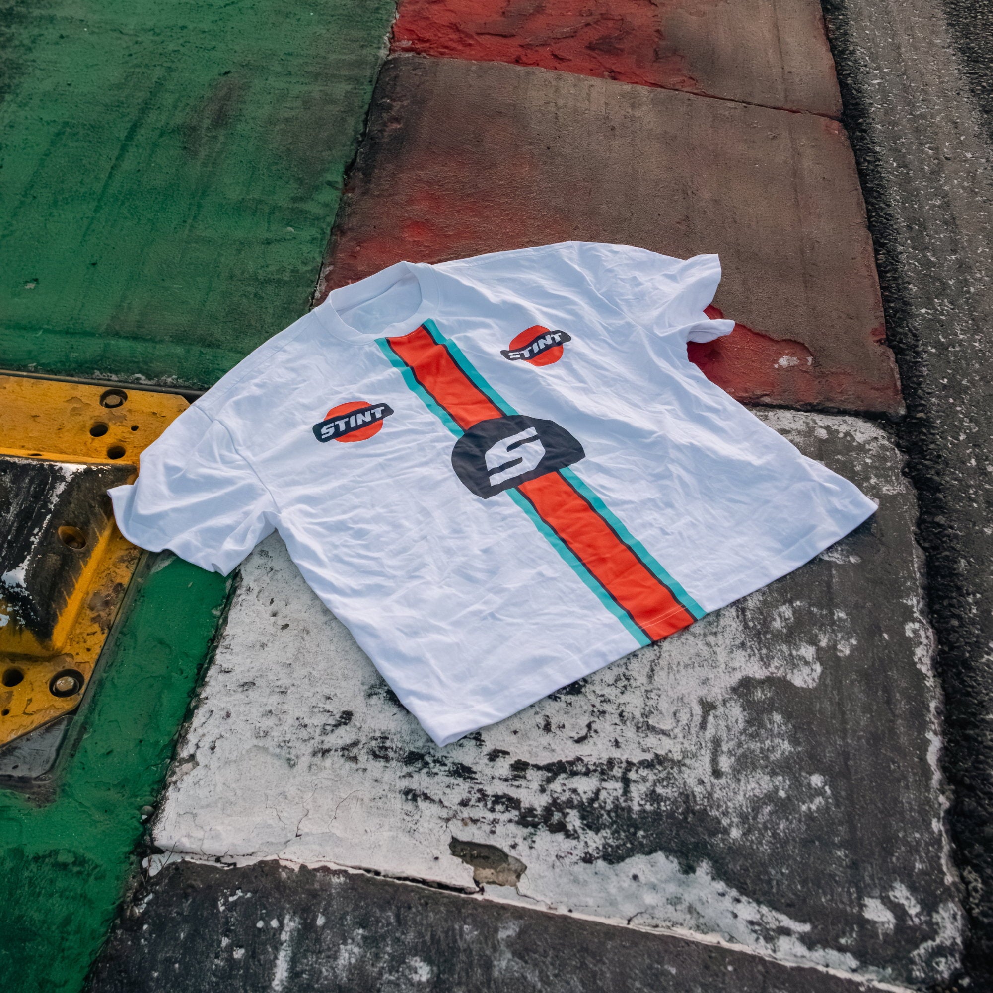 Marigold Tee by STINT GEAR. Marigold band on white, laid on green and red painted kerbing. Motorsport livery apparel.