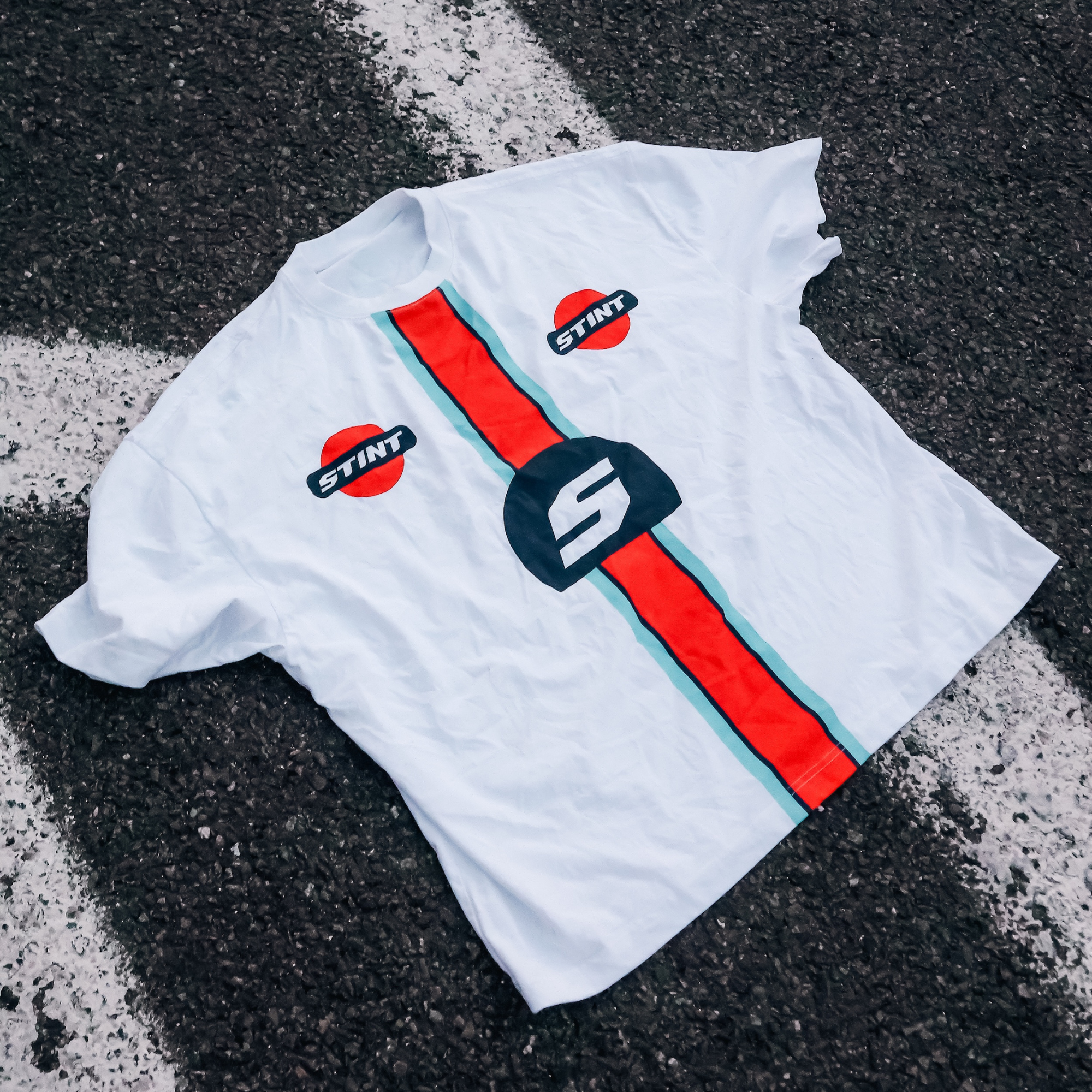 Marigold Tee by STINT GEAR. Marigold band on white, laid on painted track asphalt. Motorsport livery apparel.