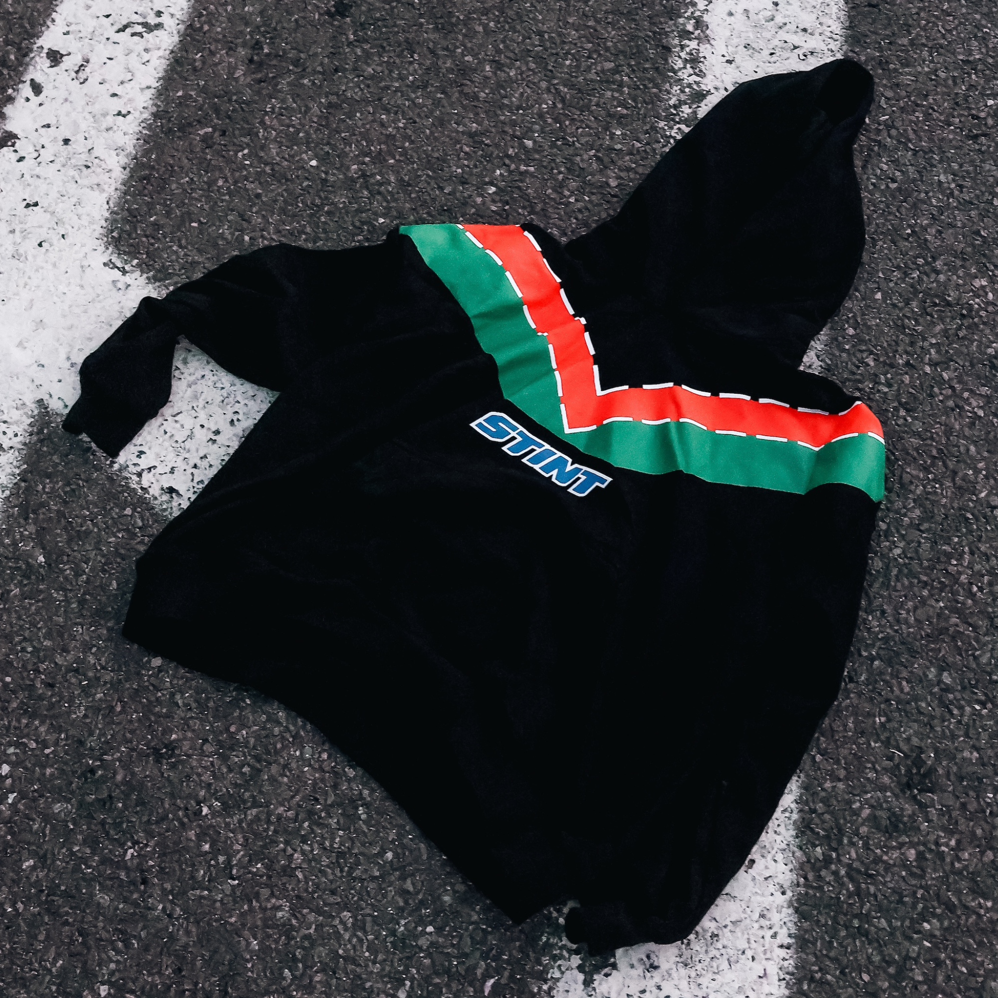 Rotary Hoodie by STINT GEAR. Orange and green 787B livery on black, laid on painted track asphalt. Motorsport livery apparel.