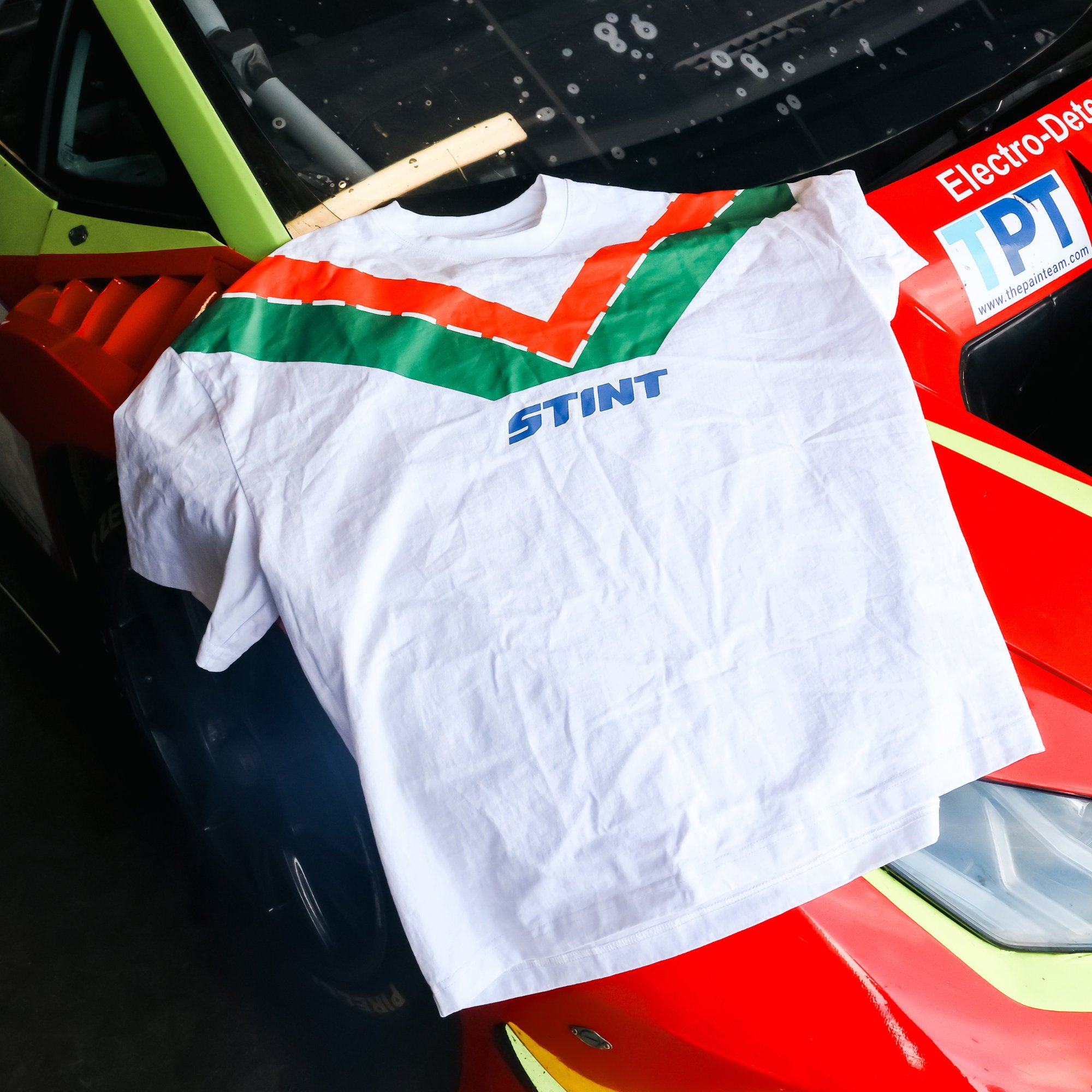 Rotary Tee by STINT GEAR. Orange and green 787B livery on white, over the nose of a race car in a garage. Motorsport livery apparel.