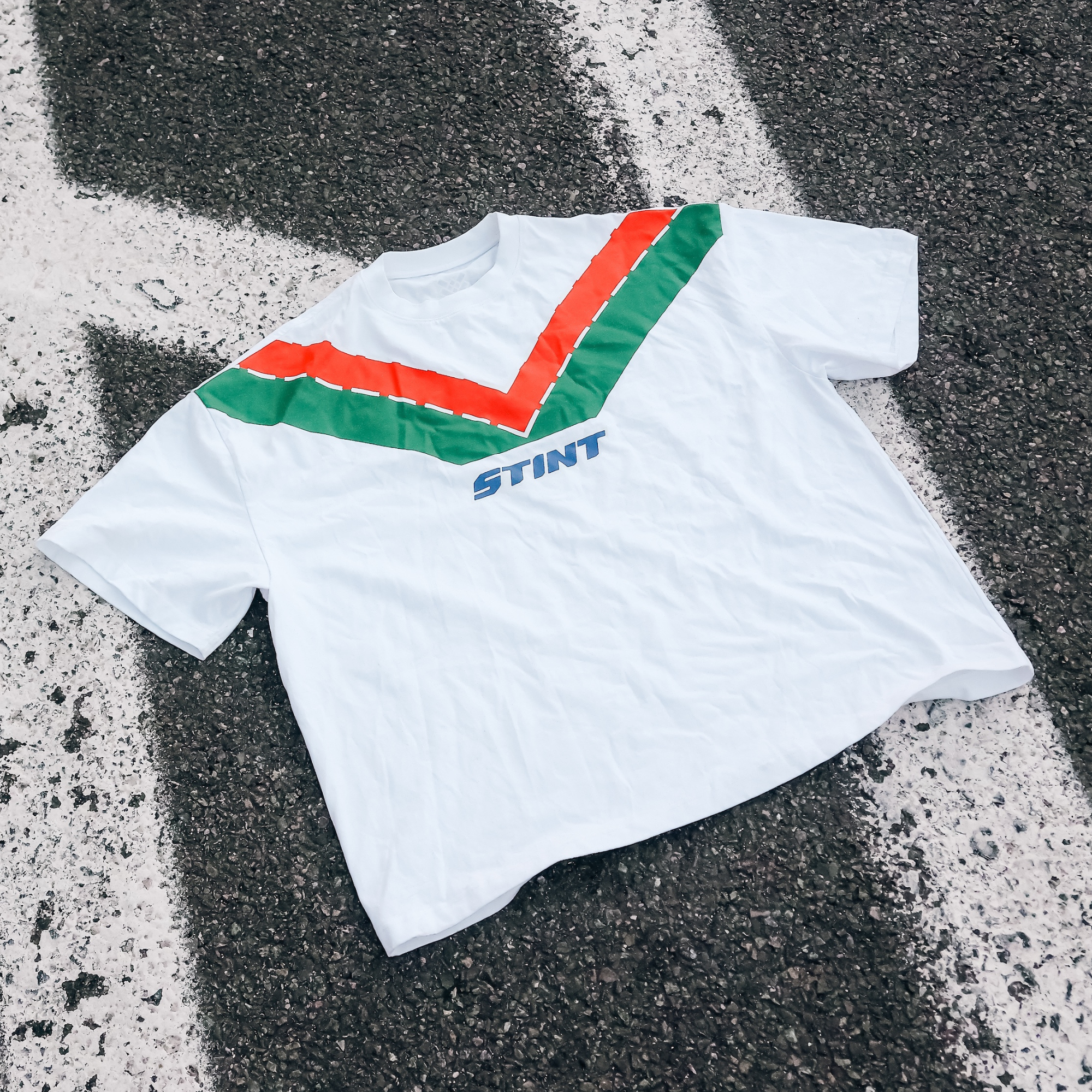 Rotary Tee by STINT GEAR. Orange and green 787B livery on white, laid on painted track asphalt. Motorsport livery apparel.